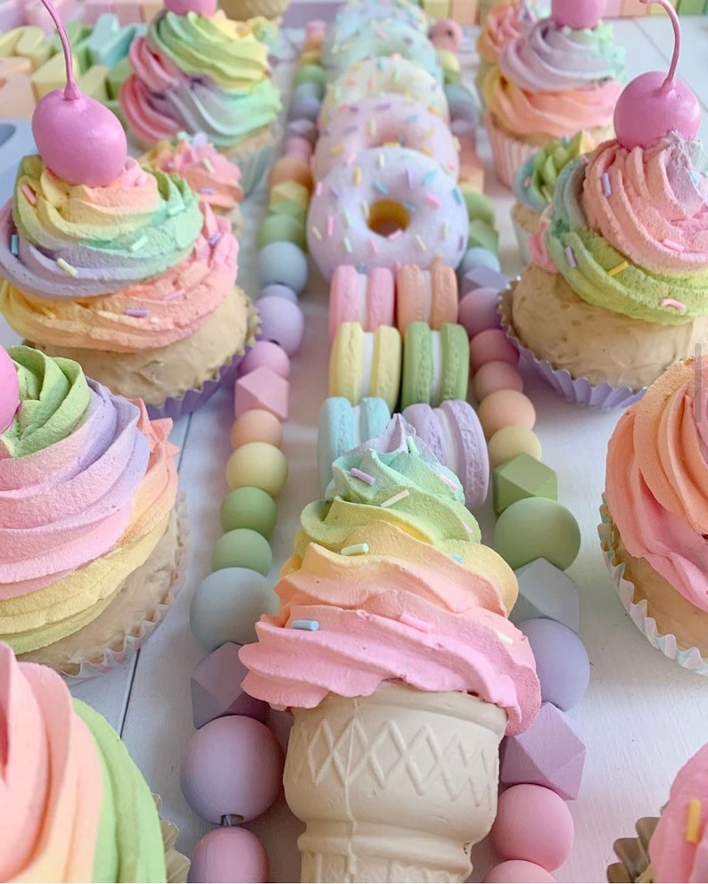 Puede incluir: Cupcakes, donuts y un cono de helado de color pastel con chispas, dispuestos sobre una superficie blanca con una guirnalda de cuentas de color pastel.