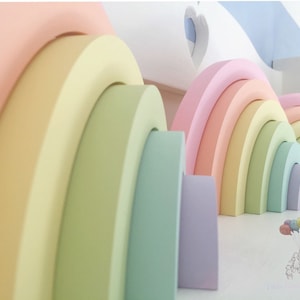 May include: A pastel rainbow arch made of wood with rounded edges. The arch is made up of seven different colours: pink, yellow, green, light blue, purple, light green, and orange. The arch is stacked in a way that the largest piece is at the bottom and the smallest piece is at the top.