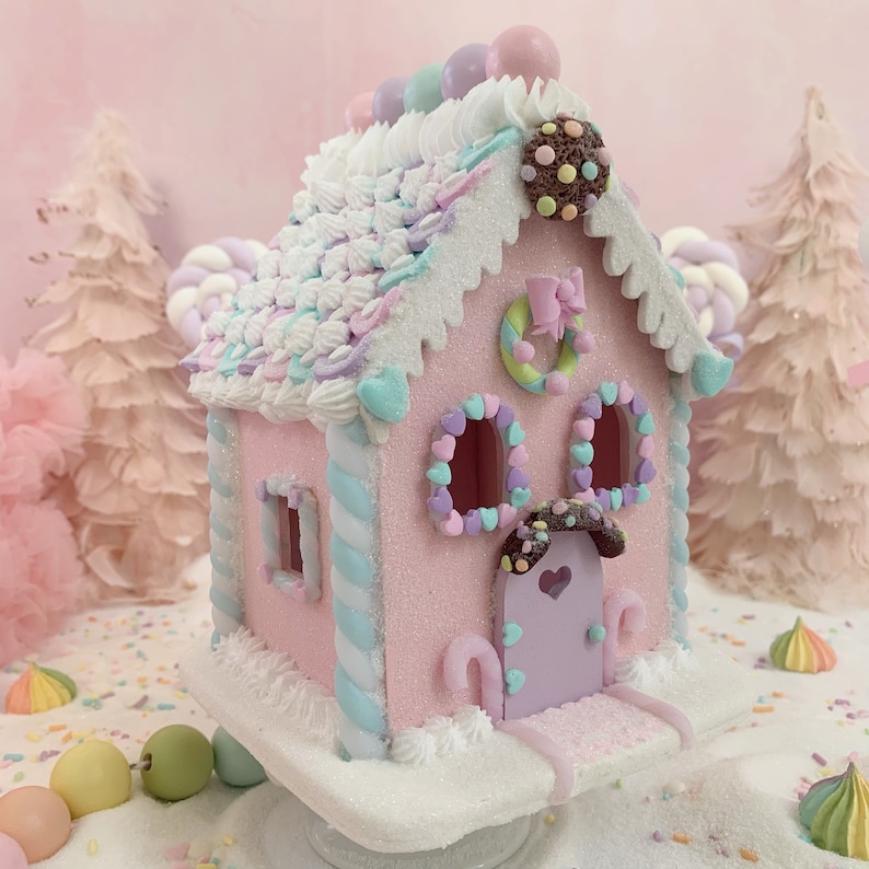 K&ouml;nnte beinhalten: Ein rosa Lebkuchenhaus mit wei&szlig;em Zuckerguss, Pastellbonbons und dekorativen Akzenten. Das Dach ist mit wei&szlig;em Zuckerguss und Pastellstreuseln bedeckt. Das Haus hat herzf&ouml;rmige Fensterdekorationen und eine lavendelfarbene T&uuml;r.