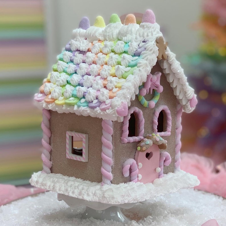 K&ouml;nnte beinhalten: Ein skurriles Lebkuchenhaus mit einem Regenbogen-Dach und Pastellakzenten. Das Haus hat eine rosa T&uuml;r, Zuckerstangen-Details und einen dekorativen Kranz. Es steht auf einem wei&szlig;en Sockel und schafft eine festliche und dekorative Pr&auml;sentation.