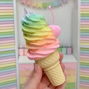 Puede incluir: Un cono de helado de colores arco&iacute;ris con una cereza rosa en la parte superior. El cono est&aacute; sostenido en una mano y est&aacute; hecho de un material amarillo claro. El helado es un remolino de rosa, amarillo, verde y azul.