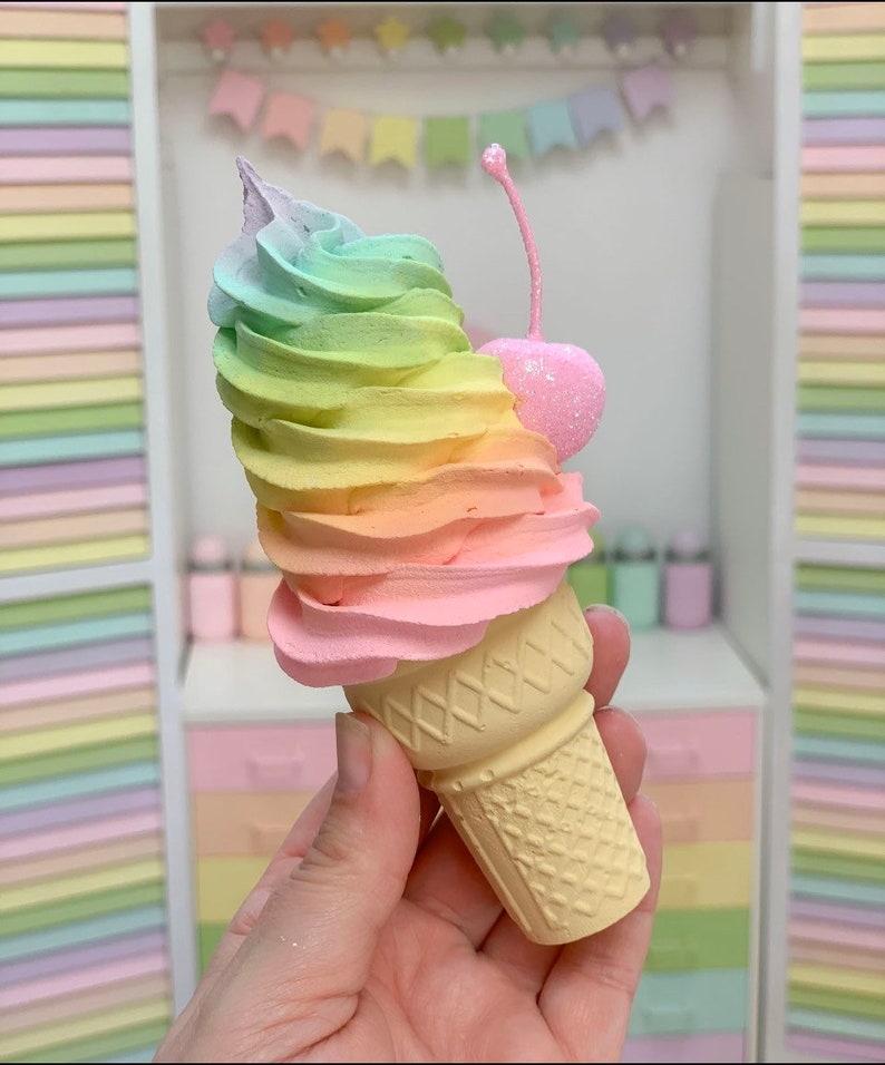 Puede incluir: Un cono de helado de colores arco&iacute;ris con una cereza rosa en la parte superior. El cono est&aacute; sostenido en una mano y est&aacute; hecho de un material amarillo claro. El helado es un remolino de rosa, amarillo, verde y azul.