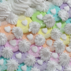 K&ouml;nnte beinhalten: Nahaufnahme eines farbenfrohen, pastellfarbenen Dekorationsartikels. Der Artikel zeigt einen Regenbogen aus Farben, darunter Rosa, Gelb, Gr&uuml;n und Blau, mit wei&szlig;em Zuckerguss und herzf&ouml;rmigen Dekorationen. Die Oberfl&auml;che ist mit Glitzer bedeckt.