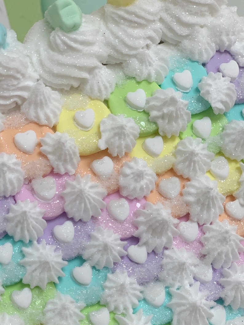 K&ouml;nnte beinhalten: Nahaufnahme eines farbenfrohen, pastellfarbenen Dekorationsartikels. Der Artikel zeigt einen Regenbogen aus Farben, darunter Rosa, Gelb, Gr&uuml;n und Blau, mit wei&szlig;em Zuckerguss und herzf&ouml;rmigen Dekorationen. Die Oberfl&auml;che ist mit Glitzer bedeckt.
