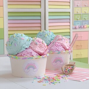May include: Two white cups with rainbow lids filled with pink and blue ice cream topped with colorful sprinkles. The cups have a sticker with a rainbow and the text "Tilda Loves Teddy".