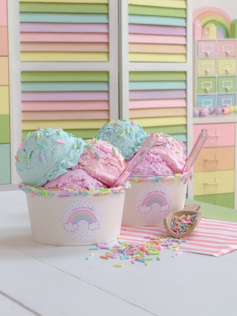 May include: Two white cups with rainbow lids filled with pink and blue ice cream topped with colorful sprinkles. The cups have a sticker with a rainbow and the text "Tilda Loves Teddy".