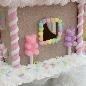 K&ouml;nnte beinhalten: Ein dekoratives Lebkuchenhaus mit glitzerndem, beigefarbenem &Auml;u&szlig;eren, verziert mit wei&szlig;em Zuckerguss, Pastellbonbons und einem herzf&ouml;rmigen Fenster. Rosa und wei&szlig;e Zuckerstangen-S&auml;ulen st&uuml;tzen das Dach. Die Szene ist mit Streuseln und Gummib&auml;rchen dekoriert.