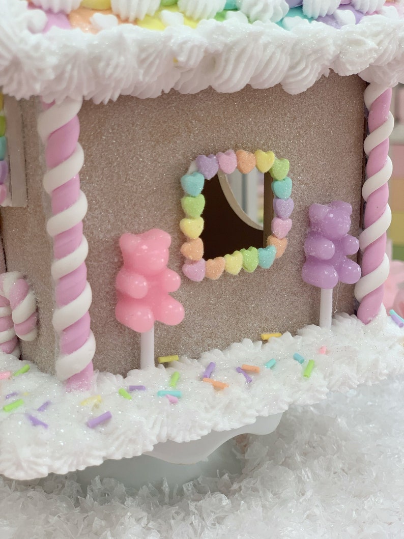 K&ouml;nnte beinhalten: Ein dekoratives Lebkuchenhaus mit glitzerndem, beigefarbenem &Auml;u&szlig;eren, verziert mit wei&szlig;em Zuckerguss, Pastellbonbons und einem herzf&ouml;rmigen Fenster. Rosa und wei&szlig;e Zuckerstangen-S&auml;ulen st&uuml;tzen das Dach. Die Szene ist mit Streuseln und Gummib&auml;rchen dekoriert.