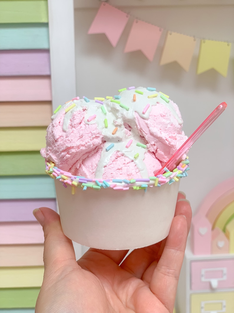 May include: A pink and white ice cream sundae with sprinkles in a white paper cup. The ice cream is topped with white frosting and sprinkles.