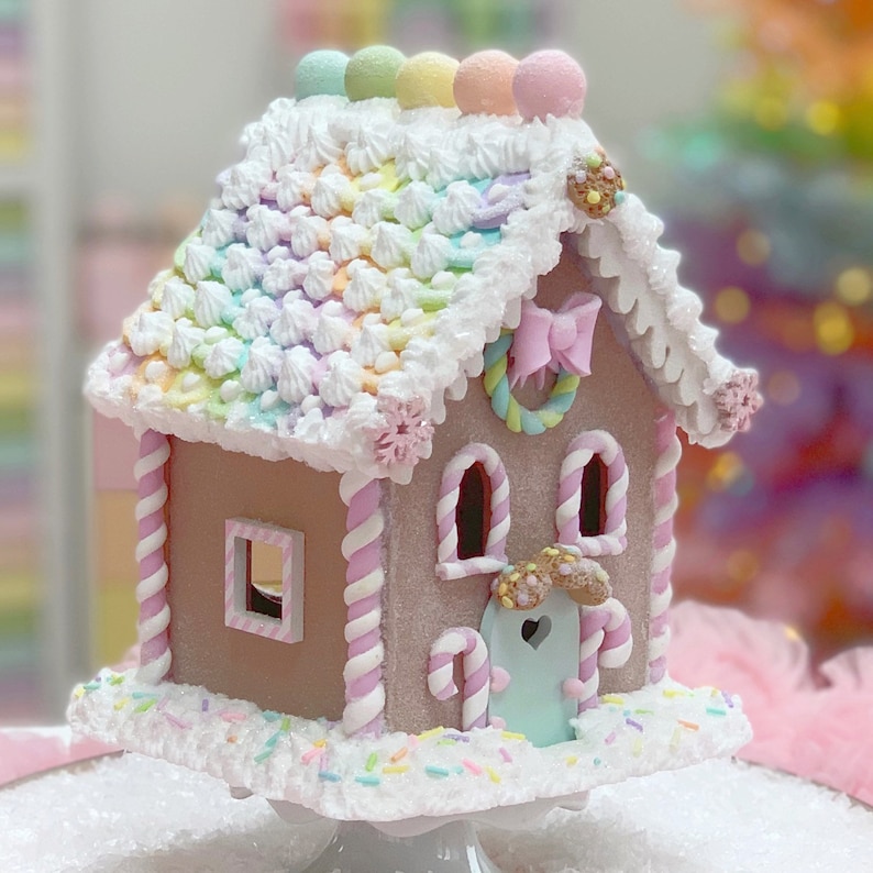 K&ouml;nnte beinhalten: Ein skurriles Lebkuchenhaus mit einem pastellfarbenen Regenbogen-Dach, wei&szlig;em Zuckerguss und Zuckerstangen-Akzenten. Das Haus hat eine hellblaue T&uuml;r, eine rosa Schleife und einen dekorativen Kranz. Es steht auf einem wei&szlig;en Sockel, der mit bunten Streuseln bestreut ist.