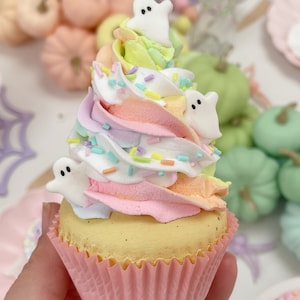 May include: A pink and yellow cupcake with rainbow frosting and white ghost sprinkles. The cupcake is topped with three white ghost candies.