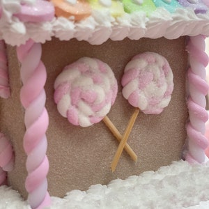K&ouml;nnte beinhalten: Ein skurriles Lebkuchenhaus mit einem Regenbogenfarbenen Dach und wei&szlig;en Zuckergussakzenten. Zwei rosa und wei&szlig; wirbelnde Lutscher sind am Haus befestigt. Das Haus ist mit rosa und wei&szlig;en, gedrehten S&uuml;&szlig;igkeitens&auml;ulen verziert.