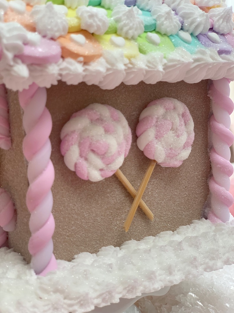 K&ouml;nnte beinhalten: Ein skurriles Lebkuchenhaus mit einem Regenbogenfarbenen Dach und wei&szlig;en Zuckergussakzenten. Zwei rosa und wei&szlig; wirbelnde Lutscher sind am Haus befestigt. Das Haus ist mit rosa und wei&szlig;en, gedrehten S&uuml;&szlig;igkeitens&auml;ulen verziert.