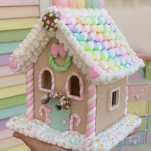 K&ouml;nnte beinhalten: Ein skurriles Lebkuchenhaus mit pastellfarbenem Zuckerguss und S&uuml;&szlig;igkeiten-Dekorationen. Das Dach ist mit regenbogenfarbenen, marshmallowartigen S&uuml;&szlig;igkeiten bedeckt. Das Haus hat rosa und wei&szlig; gedrehte S&auml;ulen, eine mintgr&uuml;ne T&uuml;r und einen Schokoladenkeks-Akzent.