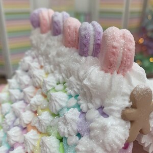 K&ouml;nnte beinhalten: Ein skurriles Lebkuchenhaus in Pastellfarben mit einem Regenbogen-Dach und wei&szlig;em Zuckerguss. Das Dach ist mit bunten Makronen und einem Lebkuchenmann verziert. Das Haus ist mit Glitzer und einer Vielzahl von Pastellfarben dekoriert.