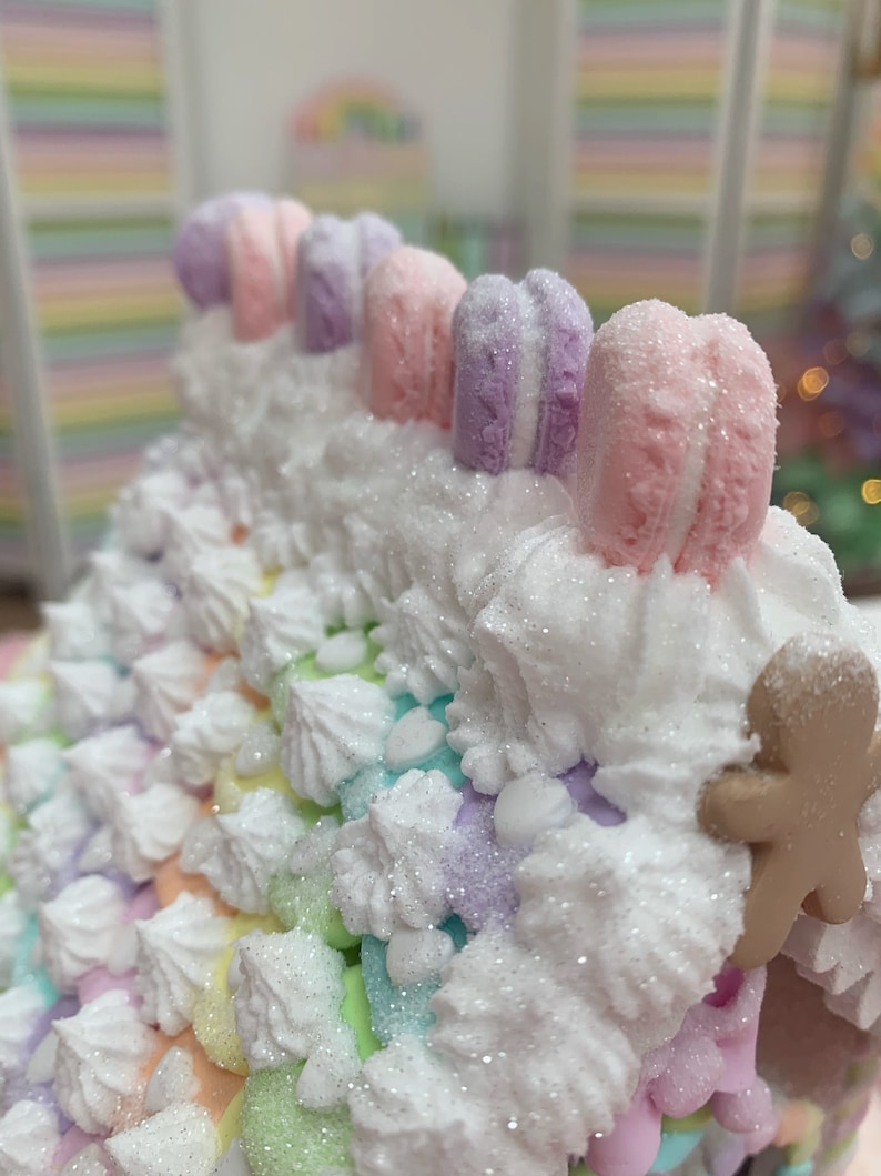 K&ouml;nnte beinhalten: Ein skurriles Lebkuchenhaus in Pastellfarben mit einem Regenbogen-Dach und wei&szlig;em Zuckerguss. Das Dach ist mit bunten Makronen und einem Lebkuchenmann verziert. Das Haus ist mit Glitzer und einer Vielzahl von Pastellfarben dekoriert.