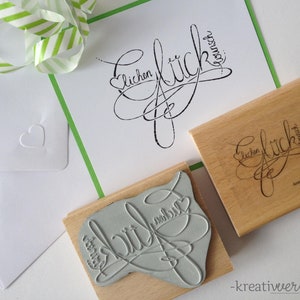 May include: Two wooden stamps with the word "Gl&uuml;ck" (German for "luck") written in cursive script. The stamps are on a white surface with a green and white striped ribbon.