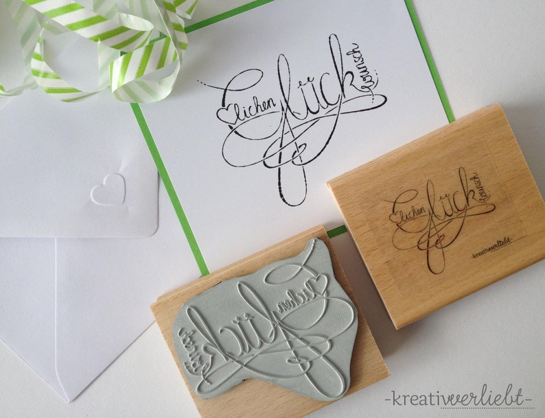 May include: Two wooden stamps with the word "Gl&uuml;ck" (German for "luck") written in cursive script. The stamps are on a white surface with a green and white striped ribbon.
