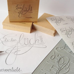 May include: A set of wooden stamps with the word "Gl&uuml;ckwunsch" (German for congratulations) in a cursive script. The stamps are used to create custom greeting cards and other paper crafts.