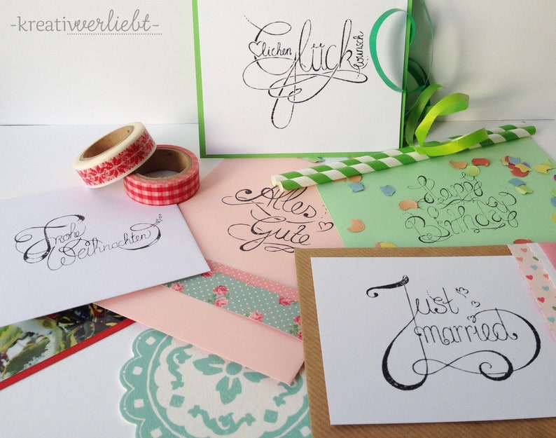 May include: Five different cards with handwritten calligraphy on them. The cards are on a white surface with colorful washi tape and a green straw. The cards say "Frohe Weihnachten", "Alles Gute", "Happy Birthday", "Just Married", and "Lieben Gl&uuml;ckwunsch".