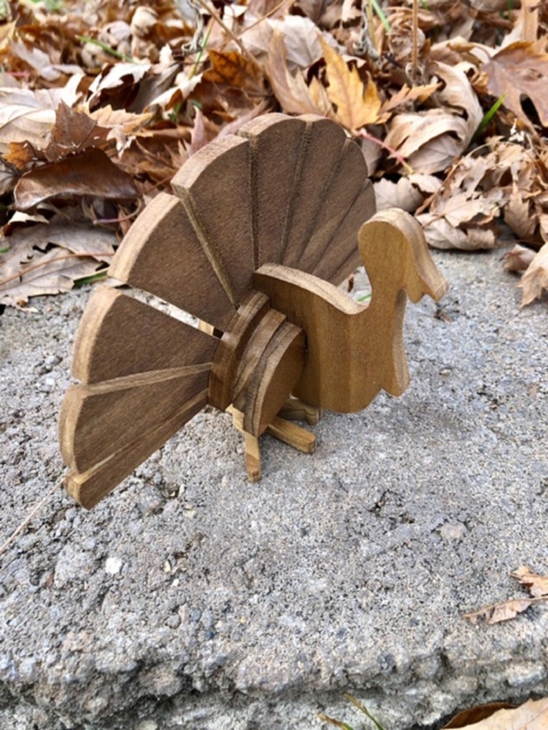 Op de afbeelding: Een houten kalkoenfiguur met een bruine afwerking. De kalkoen heeft een grote waaiervormige staart en een klein hoofd met een snavel. Het figuur staat op een betonnen oppervlak.