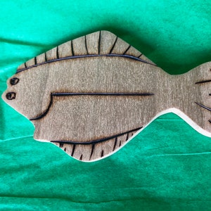 May include: A wooden cutout of a brown fish with black lines. The fish is flat and has a slightly curved shape.