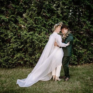 May include: Two people in wedding attire stand in front of a green hedge. The person on the left is wearing a white jumpsuit with a long, flowing veil. The person on the right is wearing a dark green suit. They are embracing and looking at each other.
