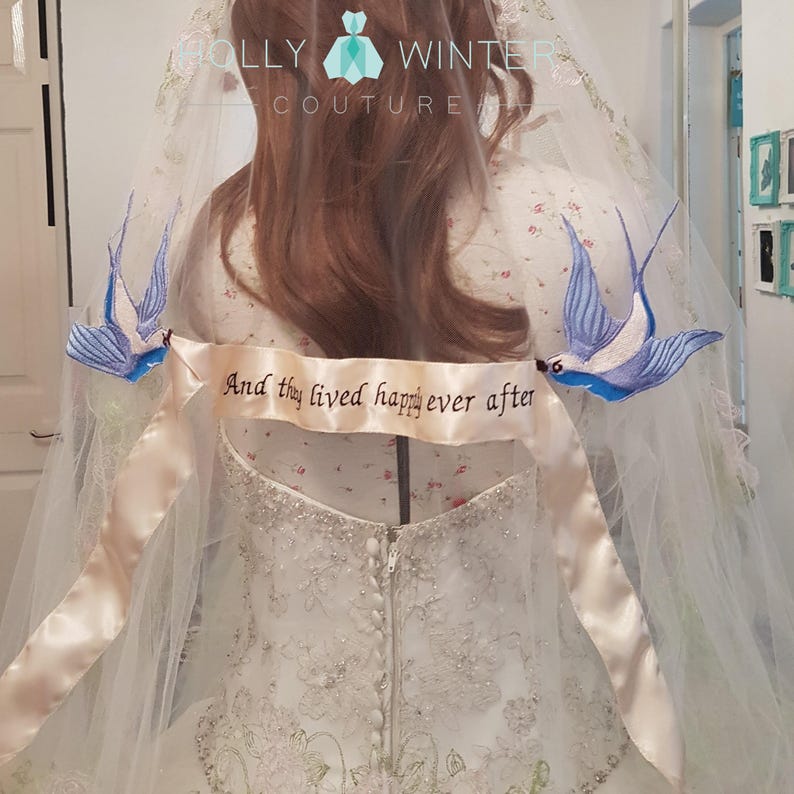 May include: A white wedding veil with a satin ribbon that reads "And they lived happily ever after". Two blue embroidered birds are attached to the veil.