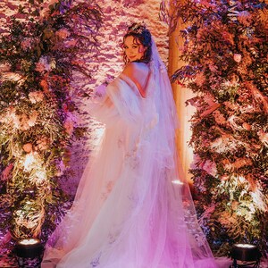 May include: A bride wearing a white tulle wedding dress with floral embroidery and a veil. She is standing in front of a wall with pink and purple lighting and floral arrangements.