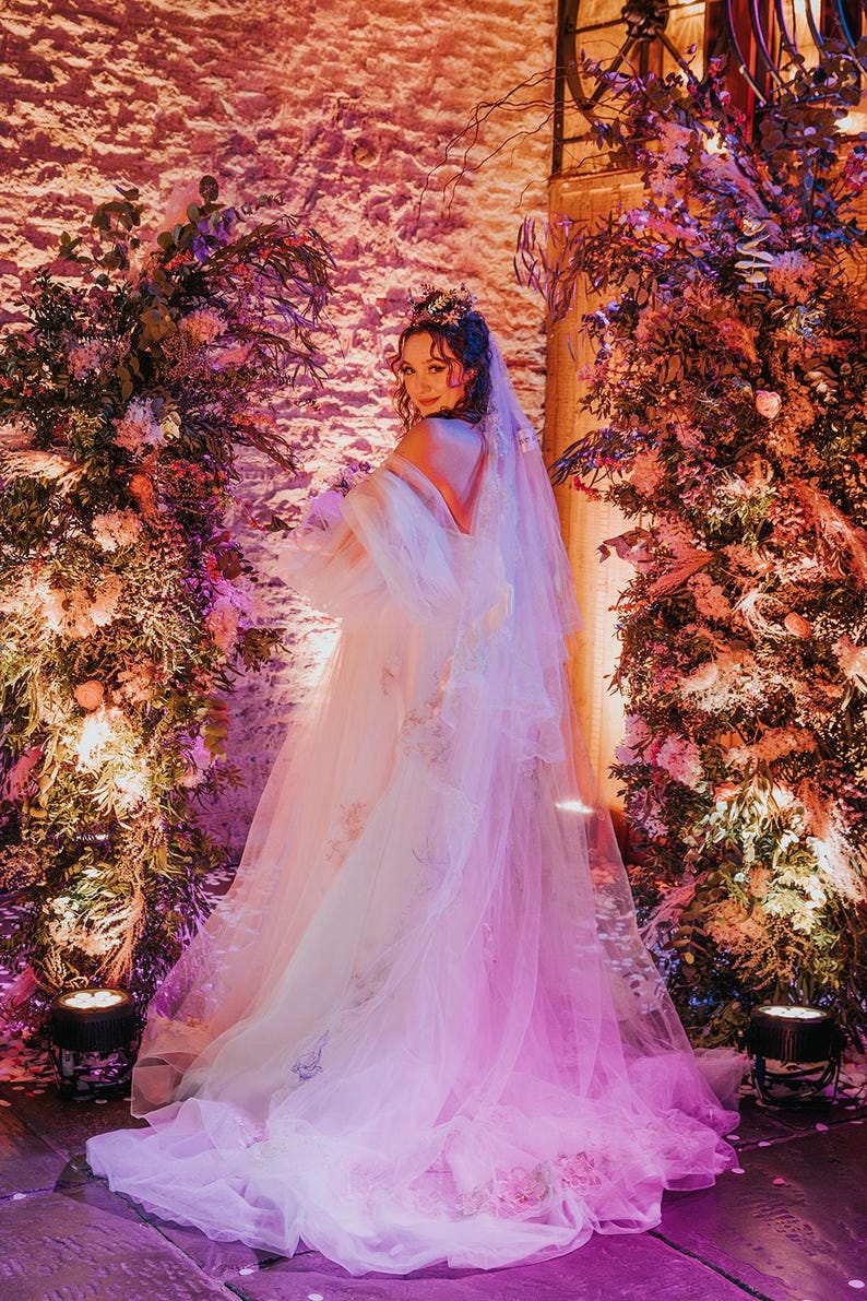 May include: A bride wearing a white tulle wedding dress with floral embroidery and a veil. She is standing in front of a wall with pink and purple lighting and floral arrangements.
