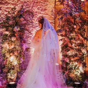 May include: A bride wearing a white wedding dress with a long, flowing veil. The dress is decorated with delicate embroidery and the veil is adorned with sparkling sequins. The bride is standing in front of a wall decorated with lush greenery and flowers.