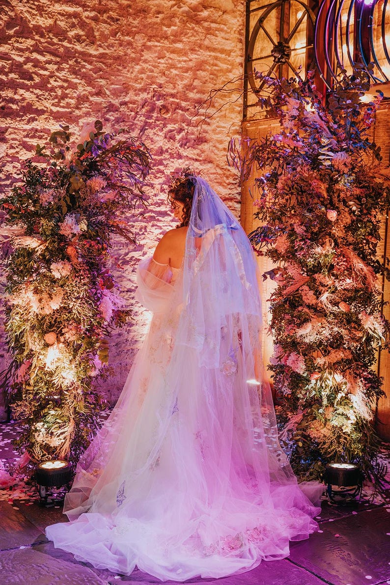 May include: A bride wearing a white wedding dress with a long, flowing veil. The dress is decorated with delicate embroidery and the veil is adorned with sparkling sequins. The bride is standing in front of a wall decorated with lush greenery and flowers.