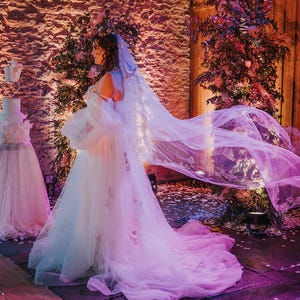 May include: A bride in a white wedding dress with a long veil, standing in a rustic setting with a white wedding cake in the background. The veil is flowing in the wind, and the bride is looking to the side.