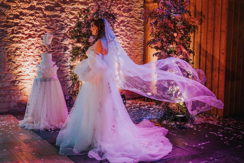 May include: A bride in a white wedding dress with a long veil, standing in a rustic setting with a white wedding cake in the background. The veil is flowing in the wind, and the bride is looking to the side.