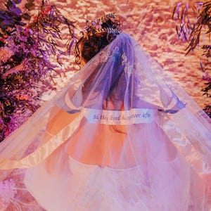 May include: A white bridal veil with delicate floral embroidery and a satin ribbon. The veil has a quote written on it: "And they lived he never after".