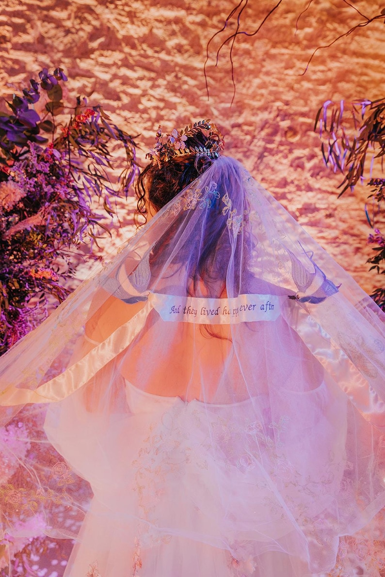 May include: A white bridal veil with delicate floral embroidery and a satin ribbon. The veil has a quote written on it: "And they lived he never after".