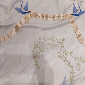 May include: A white satin fabric with embroidered floral designs and two blue birds with a ribbon that reads "Say the word and I'll be there for you".