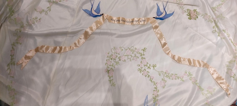 May include: A white satin fabric with embroidered floral designs and two blue birds with a ribbon that reads "Say the word and I'll be there for you".