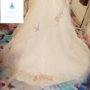 May include: A white wedding dress with delicate floral embroidery and blue birds. The dress has a full skirt and a train.