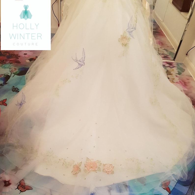 May include: A white wedding dress with delicate floral embroidery and blue birds. The dress has a full skirt and a train.