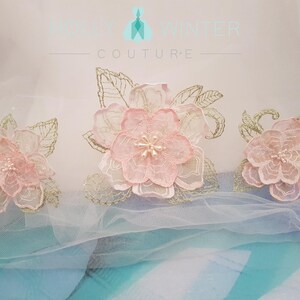 May include: Three delicate pink and gold floral appliques with pearl accents on a white tulle background. The appliques are perfect for adding a touch of elegance to any project.