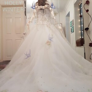 May include: A white wedding dress with a long train and a veil. The dress has floral embroidery and blue birds. The veil has a satin ribbon with the text "And so we live happily ever after".