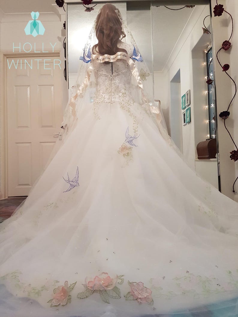 May include: A white wedding dress with a long train and a veil. The dress has floral embroidery and blue birds. The veil has a satin ribbon with the text "And so we live happily ever after".