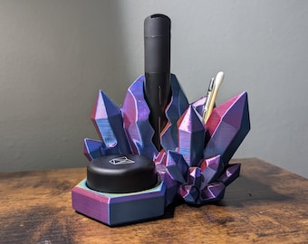 Op de afbeelding: Een veelkleurige, kristalvormige bureau-organizer. De organizer heeft een zwart cilindrisch object, een zwart cirkelvormig object en een klein wit object erin. Het kleurenschema omvat tinten paars, blauw en roze.