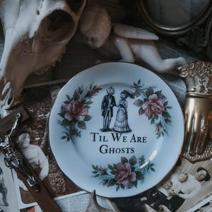 May include: A white plate with a gold rim, decorated with a couple with pumpkin heads and the text "Til We Are Ghosts." The plate is surrounded by pink roses and green leaves. The image includes a skull, a cherub, and other vintage items.