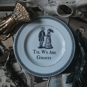 May include: A white ceramic plate with a dark blue decorative border and a central illustration of two figures with pumpkin heads, one in a suit and one in a dress holding an umbrella, with the text "TIL WE ARE GHOSTS" printed below them.