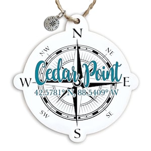 May include: White wooden compass ornament with a black compass design and the text "Cedar Point" in teal blue lettering. The coordinates "42.5781° N, 88.5409° W" are printed below the text.