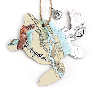 May include: A wooden sea turtle ornament with a nautical map of St. Augustine, Florida. The turtle is painted in shades of brown, green, and blue. The map is beige and blue with black lines and text. The ornament has a rope hanger and a small compass charm.