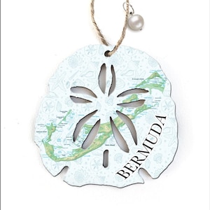 May include: A white sand dollar ornament with a map of Bermuda. The ornament has a cut-out design and is decorated with a light blue seashell pattern. It hangs from a twine string and a pearl bead.