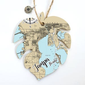 May include: A light blue and beige map of Tampa, Florida, shaped like a monstera leaf. The map includes street names and landmarks. The words "Tampa Florida" are written in black cursive script.
